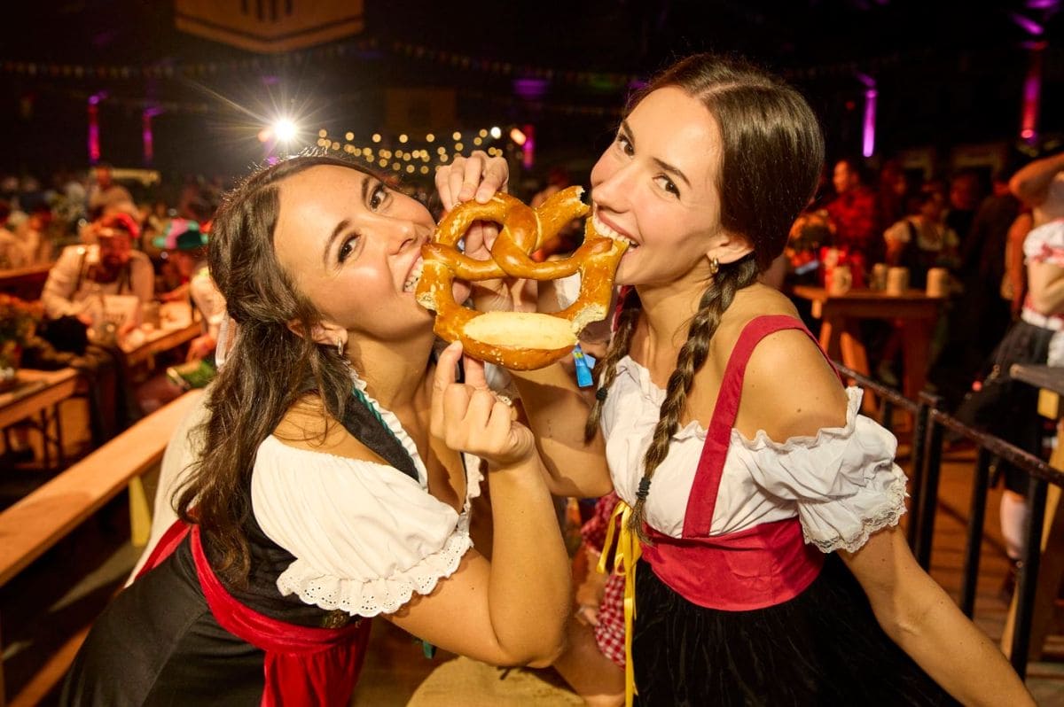 Two women in dirndl dresses sharing large pretzels at a festive beer hall or Oktoberfest-style event.",