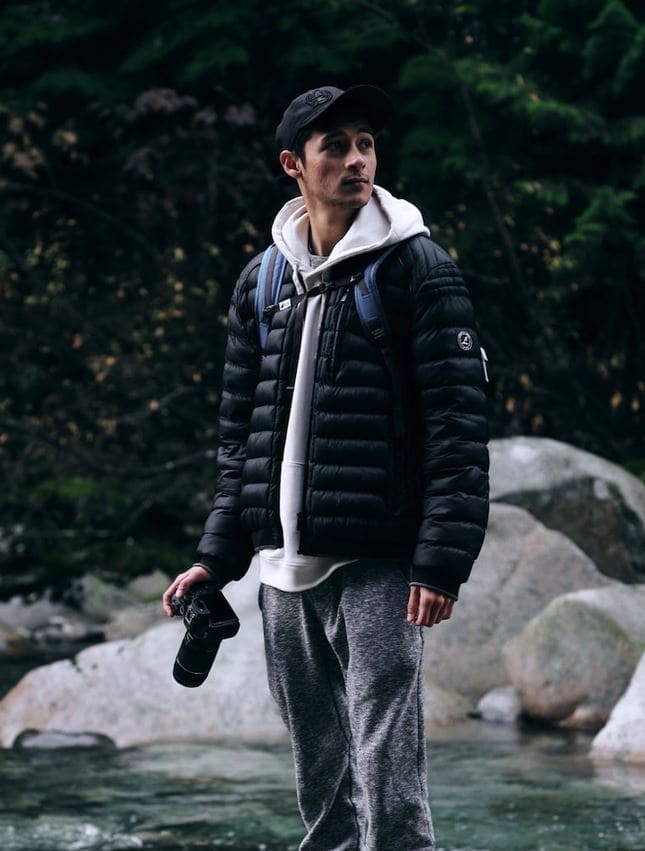 Man standing by a rocky stream in a forest, wearing a black puffer jacket over a white hoodie, holding a camera in his right hand.
