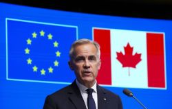 Speaker in a dark suit at a podium, with EU stars flag and Canadian flag behind on a blue backdrop.