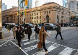 Group of pedestrians crossing a city street at a white-striped crosswalk in front of a beige historic building still in daylight.