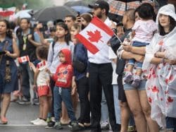 Crowd of diverse spectators at a rainy parade, many waving small Canadian flags.",