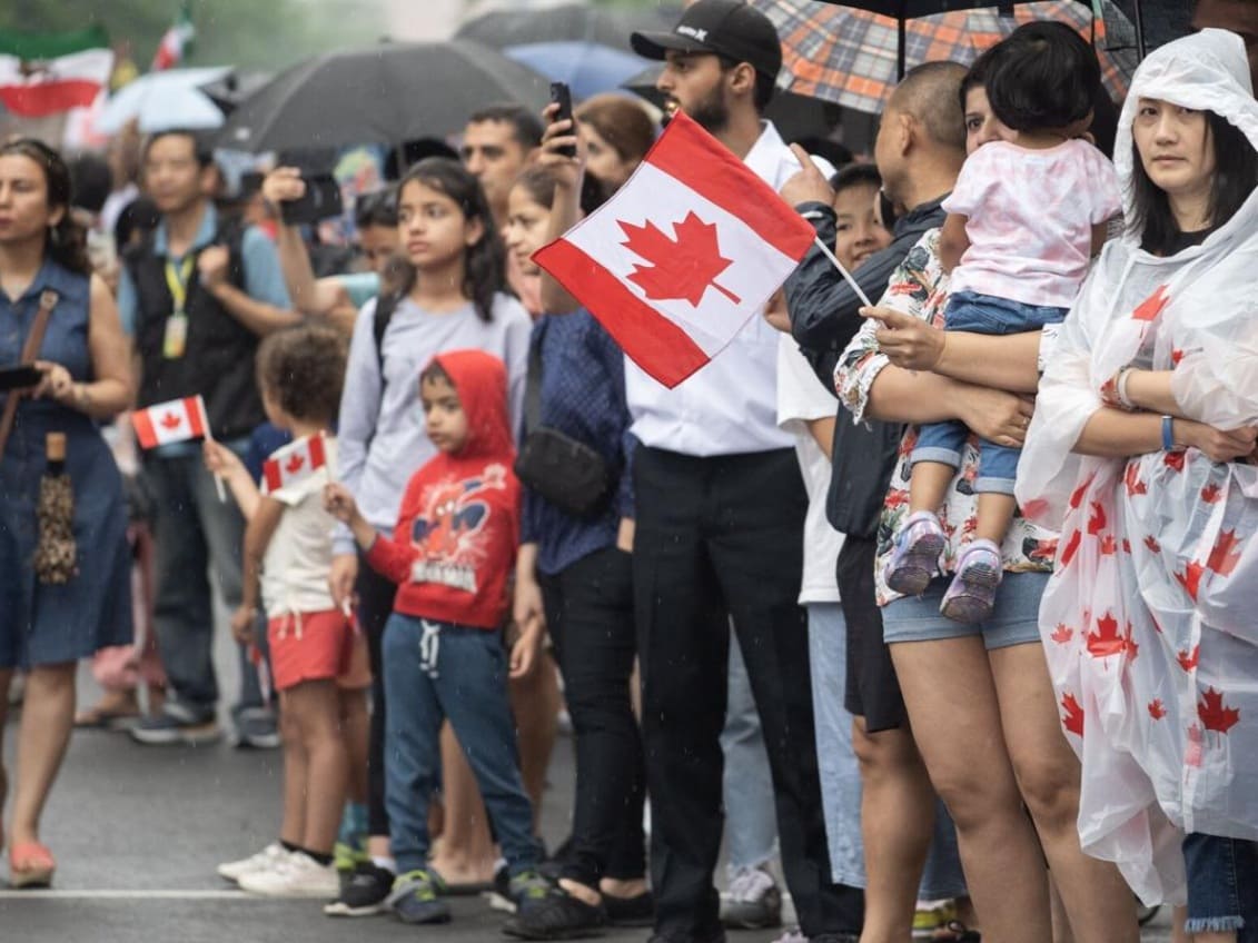 канада тммиграуия | Наш Ванкувер Crowd of diverse spectators at a rainy parade, many waving small Canadian flags.",