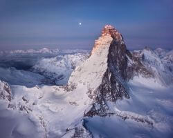 Snow-covered mountain peak under a twilight purple sky with a bright moon above the horizon, expansive glacier valleys below.