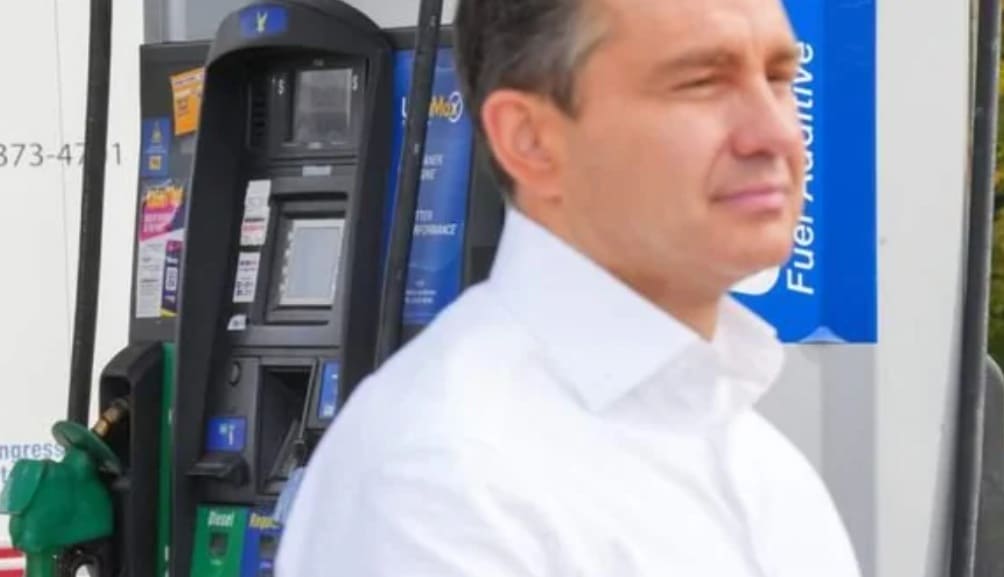 Man in a white shirt standing by a gas pump with fueling nozzle and dispenser visible in the frame.