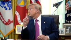 Man in a navy suit and pink tie sits at a desk, raising his index finger as if making a point, with flags and framed photos in the background.