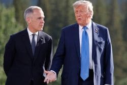 Two men in dark suits walk outdoors, talking, with a blurred green landscape behind them.