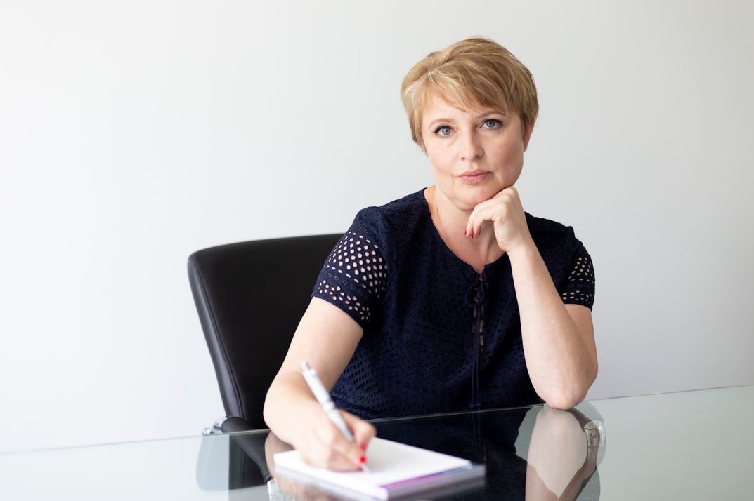 Professional woman sits at a glass desk, writing in a notebook with her hand on her chin, looking at the camera.