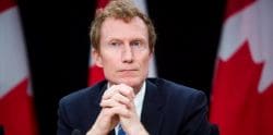 Man in a dark suit sits with hands clasped, Canadian flags visible in the background.