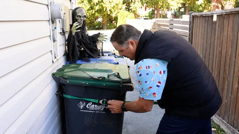 Man leaning toward a green trash can beside a house while a Halloween skeleton prop sits on the wall nearby in a narrow alleyway.
