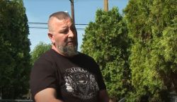 Bearded man in a black graphic T-shirt standing outdoors with green hedges and a utility pole in the background.