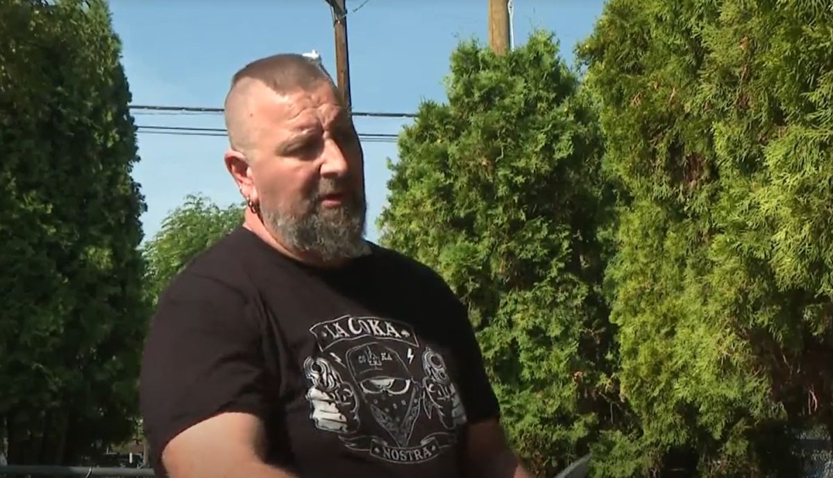 Bearded man in a black graphic T-shirt standing outdoors with green hedges and a utility pole in the background.