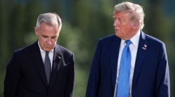 Two men in dark suits outdoors, one looking down while the other glances at him, with a blurred green background.