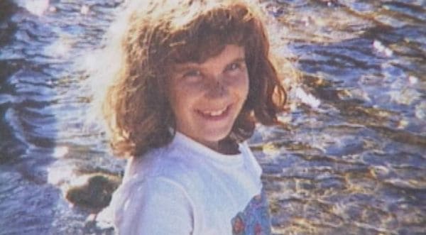 Smiling young girl with curly brown hair, wearing a white shirt, standing by sunlit water and rocks.