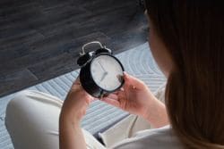 Person sitting cross-legged, holding a black analog alarm clock up close indoors.