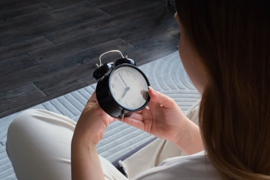 перевод часов | Наш Ванкувер Person sitting cross-legged, holding a black analog alarm clock up close indoors.