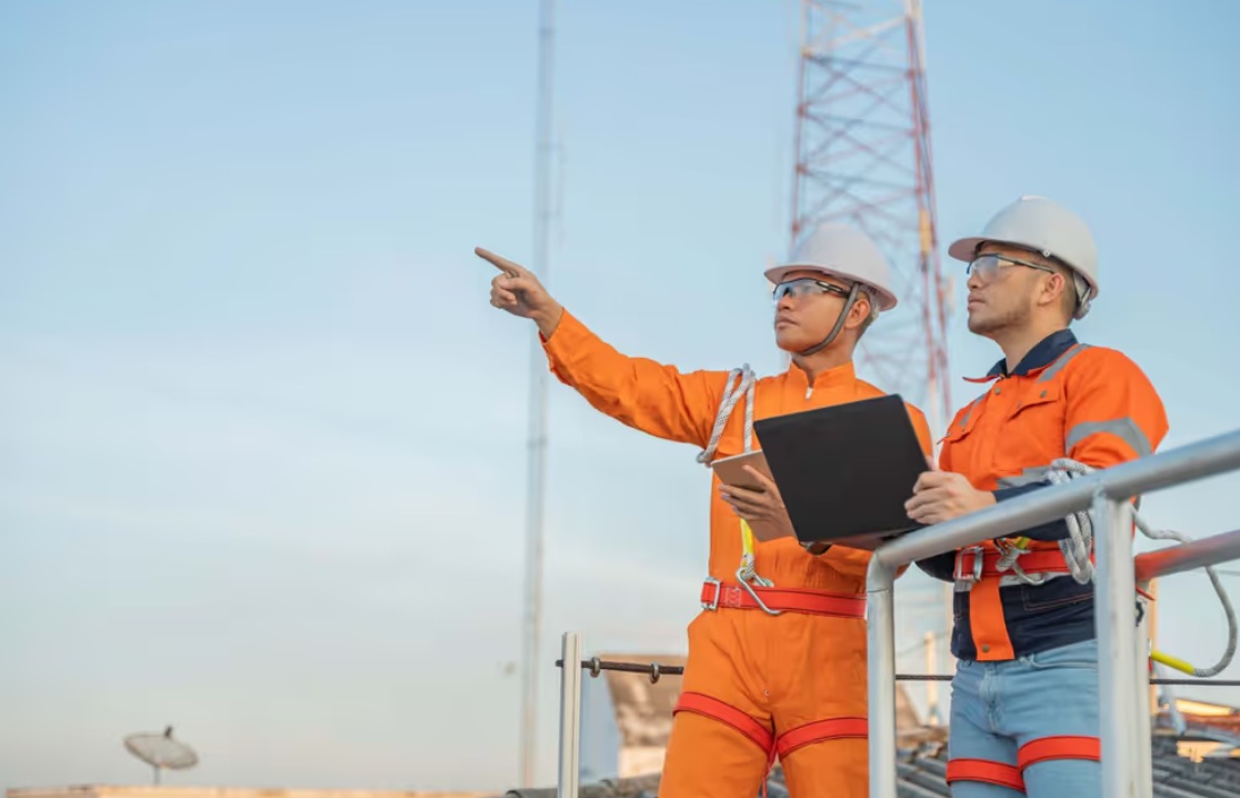повышение зарплаты | Наш Ванкувер Two construction workers in orange safety overalls and hard hats stand on a platform; one points toward something while the other holds a clipboard, with a communication tower in the backdrop.