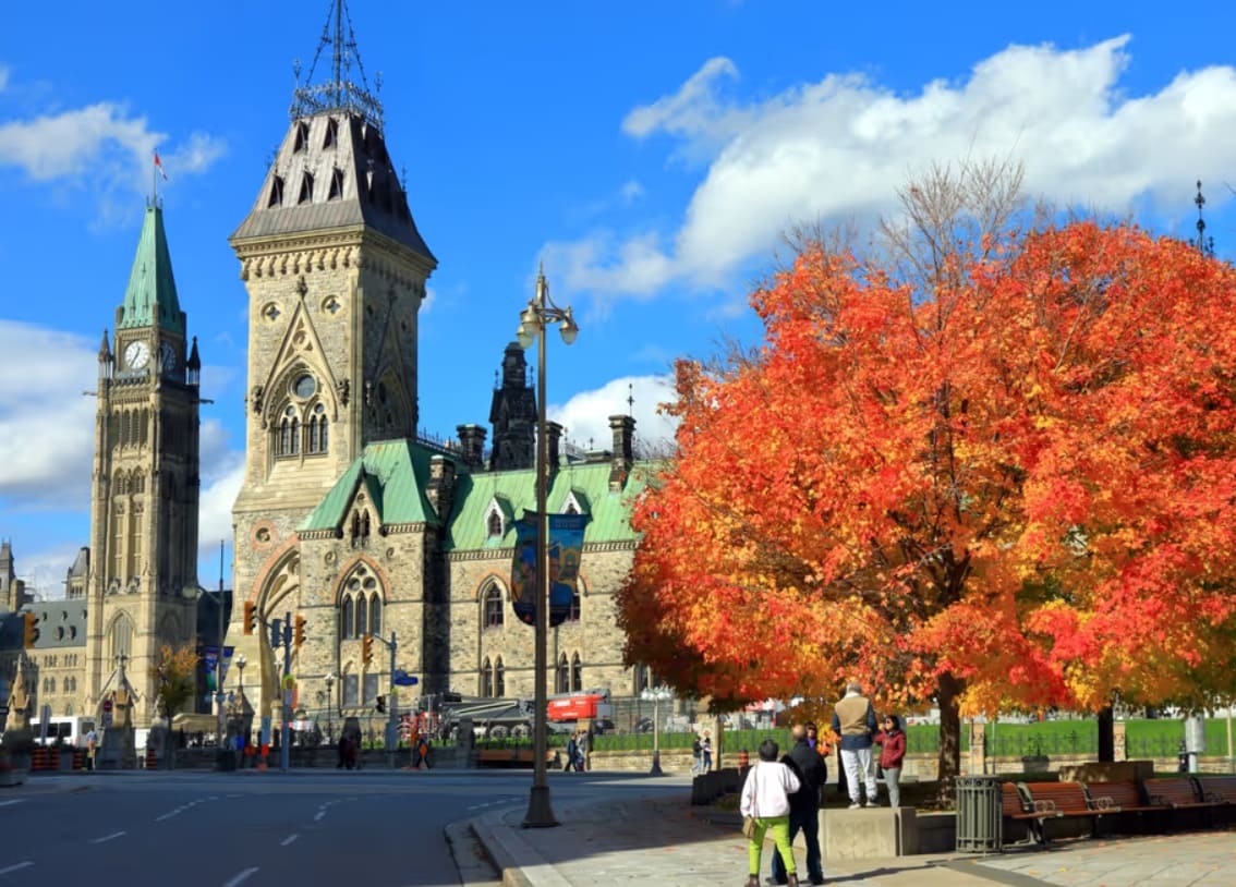 Ottawa street scene with Gothic-style Parliament buildings and bright orange fall trees in the foreground under a blue sky
