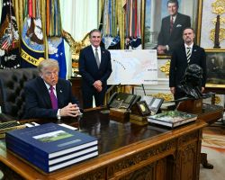 A suited man sits at an ornate wooden desk in an official office, with flags, maps, and two aides standing behind him and a bust on the desk behind.