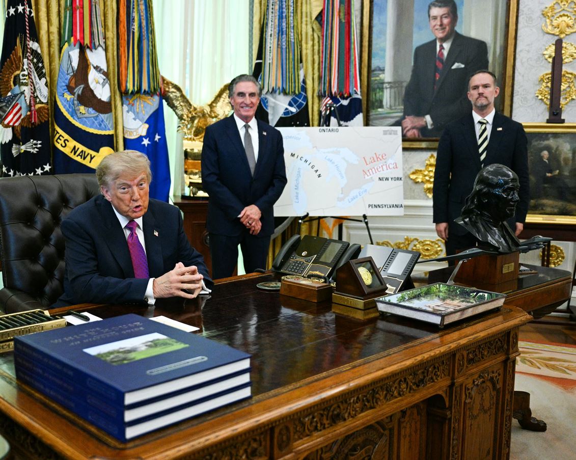 A suited man sits at an ornate wooden desk in an official office, with flags, maps, and two aides standing behind him and a bust on the desk behind.