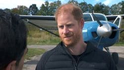 Man with red hair and beard in a dark jacket talks to someone outdoors, with a small blue propeller airplane in the background.