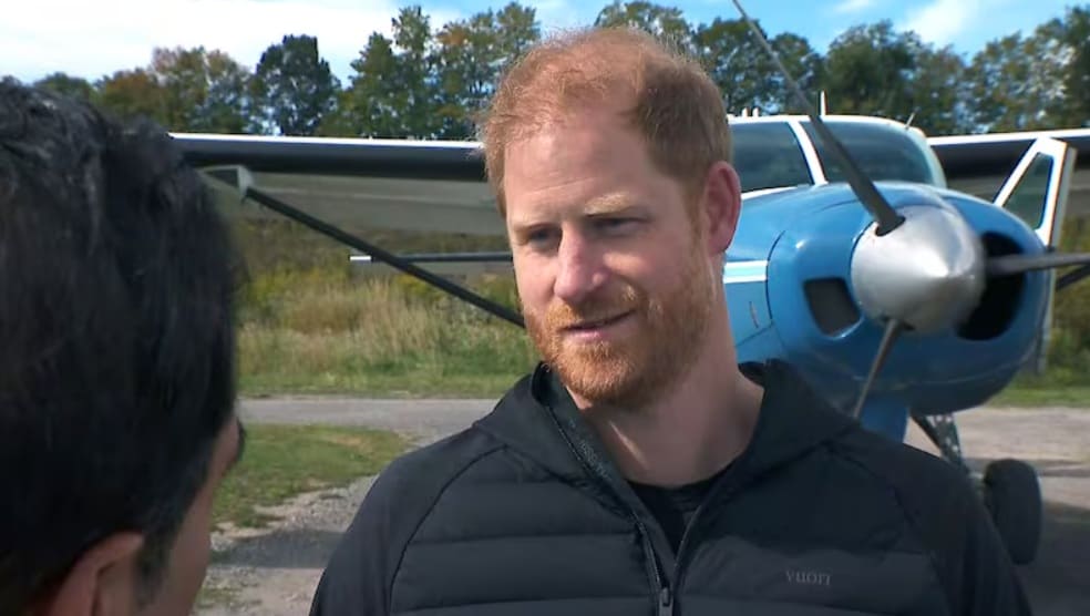 Man with red hair and beard in a dark jacket talks to someone outdoors, with a small blue propeller airplane in the background.