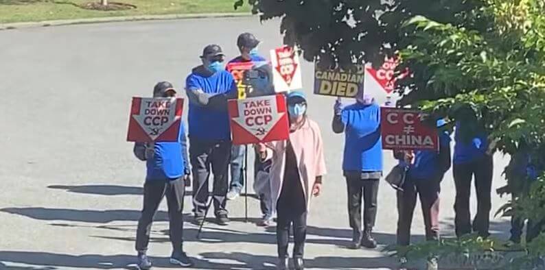 Group of protesters on a street holding red-and-white signs that say 'TAKE DOWN CCP' and other messages criticizing China/CCP, with some wear masks.