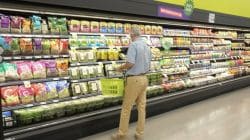 Man in a light-blue shirt and khaki pants stands in a grocery store, holding a green shopping basket while browsing bags of salad on refrigerated shelves.