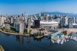 Aerial view of downtown Vancouver with high-rise buildings, the white-sailed BC Place stadium near the water, and a marina along the inlet. Mountains rise in the distance.