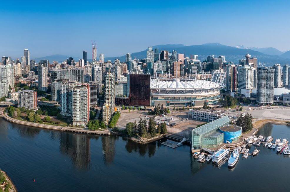 Aerial view of downtown Vancouver with high-rise buildings, the white-sailed BC Place stadium near the water, and a marina along the inlet. Mountains rise in the distance.
