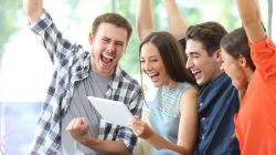 Group of five young adults cheerfully gathered around a tablet, raising their arms in celebration.
