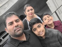 Family of four posing for a selfie outdoors, with a man in a black polo, a woman behind them, and two smiling boys nearby in front of a building and parked car.