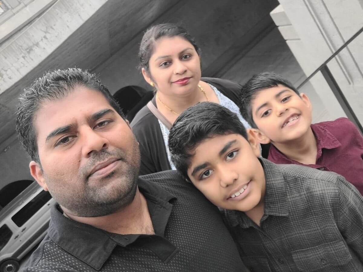 Family of four posing for a selfie outdoors, with a man in a black polo, a woman behind them, and two smiling boys nearby in front of a building and parked car.