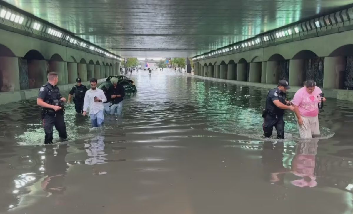 Underpass flooded with water up to mid-calf; several people wading while police assist two individuals, cars submerged in background under arched walls.