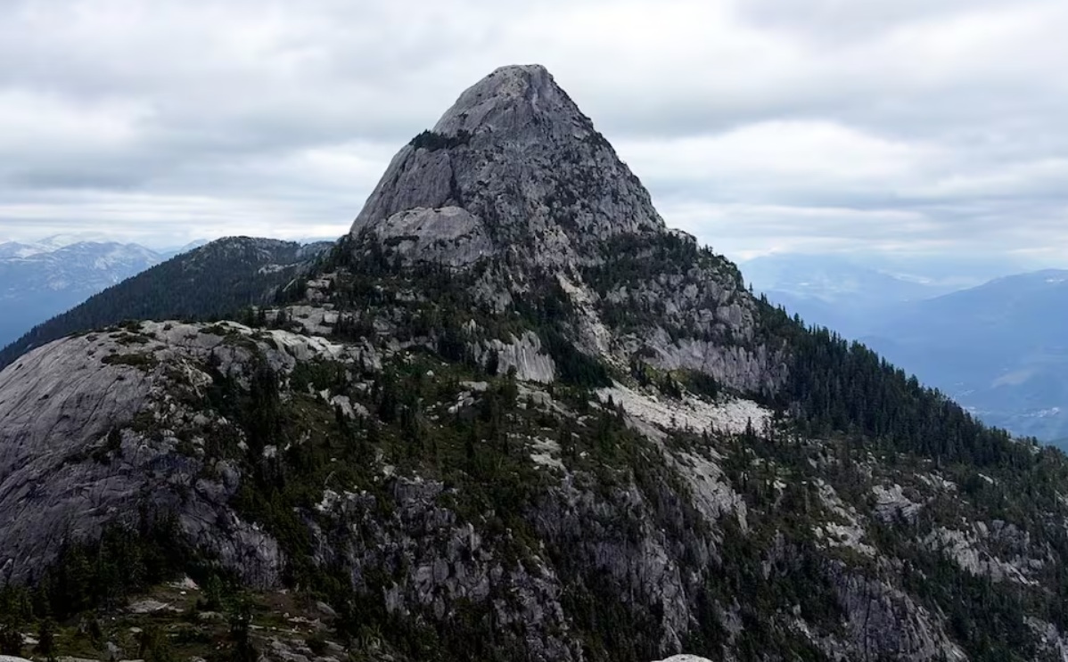 Dramatic jagged mountain peak rising above forested slopes under a cloudy sky.