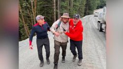 Three men walk a gravel forest road, one in a beige jacket being helped by two friends as they smile and share a moment; a truck is parked to the right.
