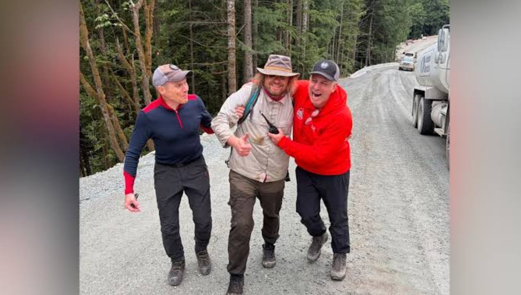 станислав воло жив | Наш Ванкувер Three men walk a gravel forest road, one in a beige jacket being helped by two friends as they smile and share a moment; a truck is parked to the right.