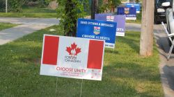 Lawn political signs on a grassy roadside, foreground sign reads Forever Canadian with a red maple leaf and 'Choose Unity'.