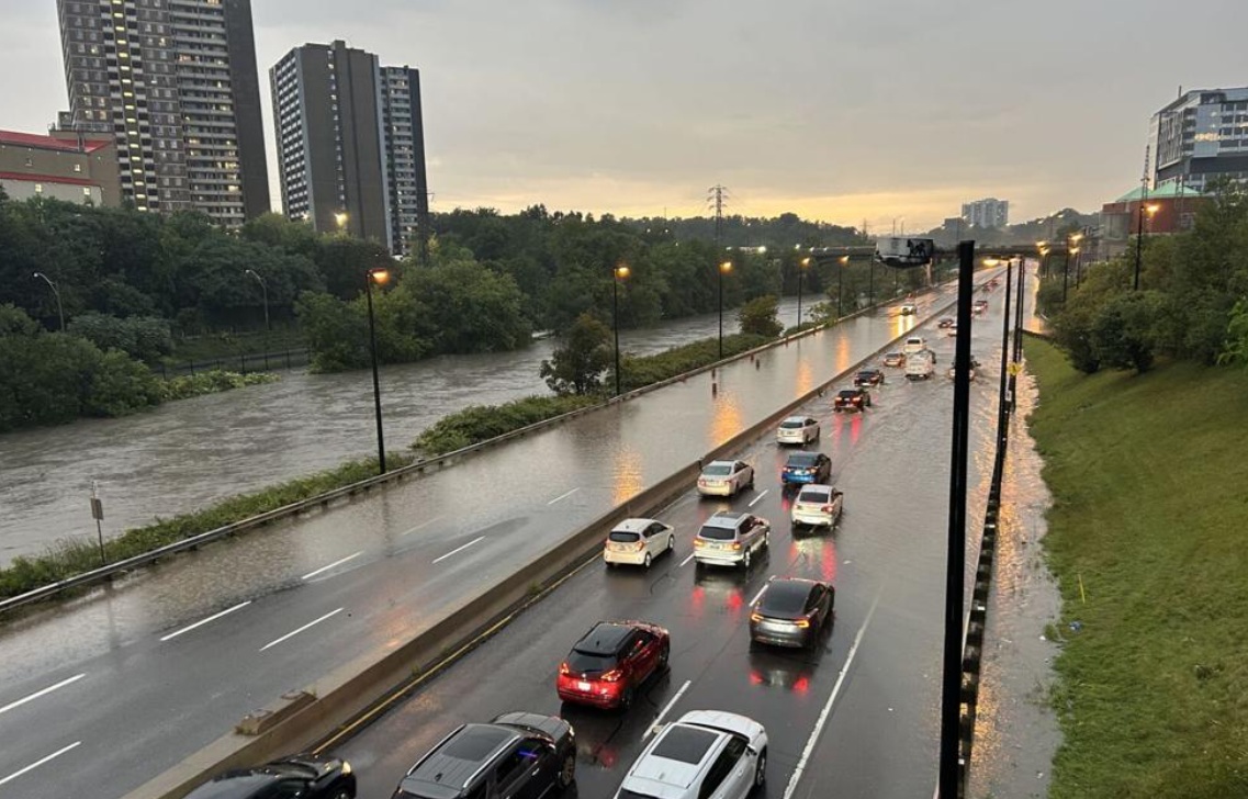Cars drive on a flooded multi-lane highway beside a swollen river, with trees and tall buildings in the background at dusk/sunset.