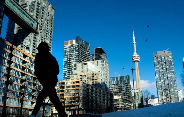 Silhouette of a person walking in the foreground with a cityscape of modern glass buildings and the CN Tower against a clear blue sky.