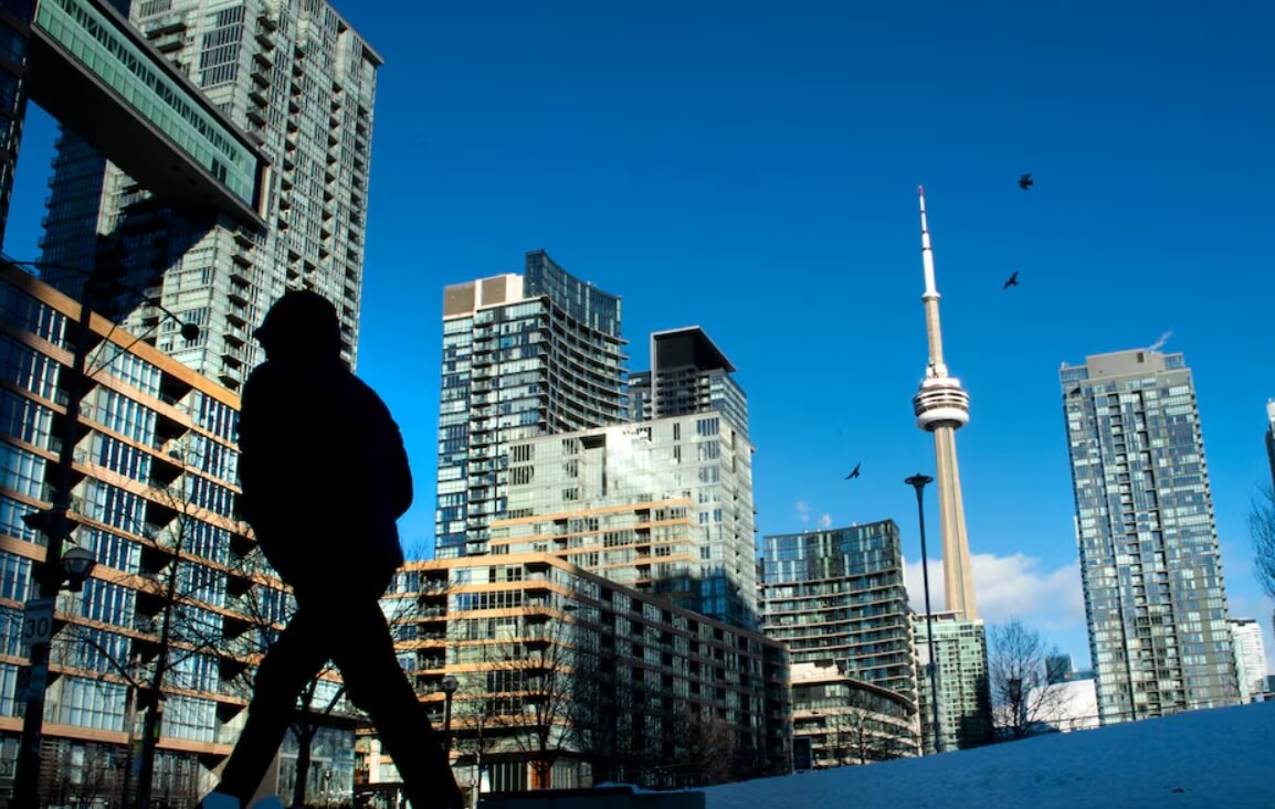торонто саммит | Наш Ванкувер Silhouette of a person walking in the foreground with a cityscape of modern glass buildings and the CN Tower against a clear blue sky.