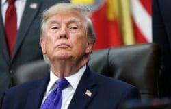 Man in a navy suit and purple tie sits in a high-backed chair with a serious expression, a flag in the background.