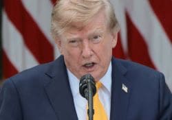 Close-up of a man in a navy suit and yellow tie speaking into a microphone, with American flags in the background.
