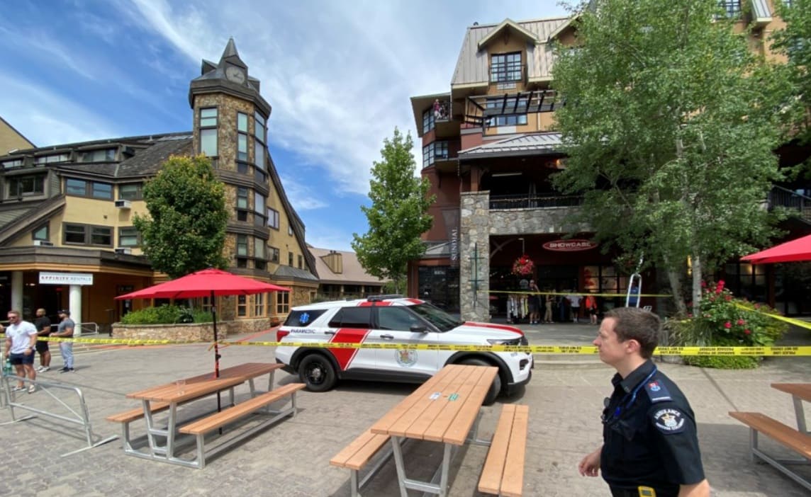 Police SUV parked in a plaza with yellow caution tape, red umbrellas, and people nearby against a backdrop of alpine-style buildings.