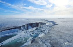 Snow-covered Arctic landscape with a deep crevasse and turquoise meltwater, extending toward the horizon under a blue sky with wispy clouds.