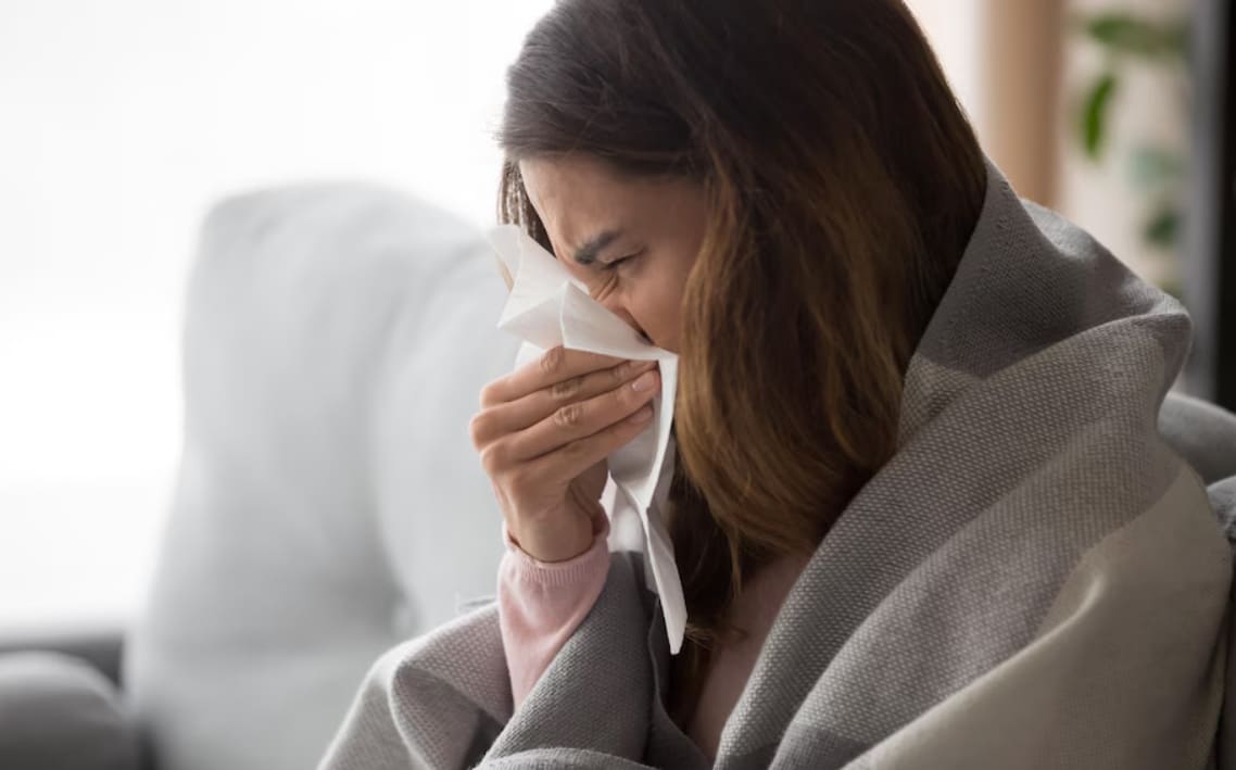 вирусы 2026 | Наш Ванкувер Woman sneezing into a tissue while wrapped in a blanket on a couch at home, eyes closed in discomfort train.