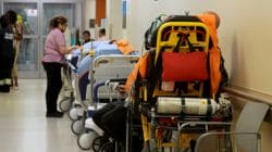 Hospital corridor with lined stretchers and medical staff tending to patients; orange emergency stretcher in the foreground.