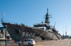 Gray naval destroyer docked at a pier with sailors on deck and hull number 3508 visible on the hull, at a sunny port.