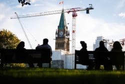 Silhouetted people sit on benches watching a clock tower under construction with cranes in the background.