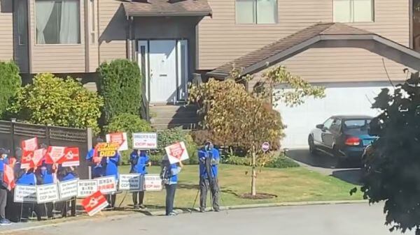 Group of protesters in blue jackets holding red-and-white signs on a residential street in front of a beige house.