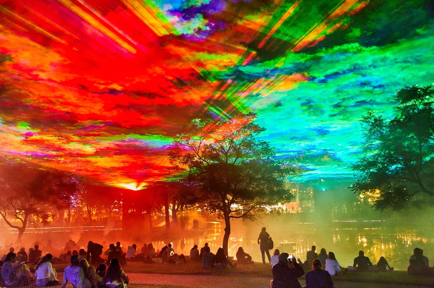 Crowd seated on the grass by a lake as a rainbow-colored light show fills the sky above trees and misty air.